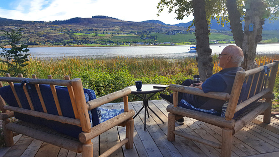 Senior man 60's/ 70's sitting by lake and wetlands enjoying his surroundings. Coffee Thermos in Hand.  Digital tablet and coffee mug on table.  Two rough hewn lounge sofas. Autumn. 