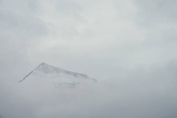 istock Minimal mountain landscape with high pointy rock in clouds. Minimalist mountain scenery with sharp snowy mountain peak over clouds. Snow-white pointed pinnacle above white clouds. Big top in dense fog 1350320167
