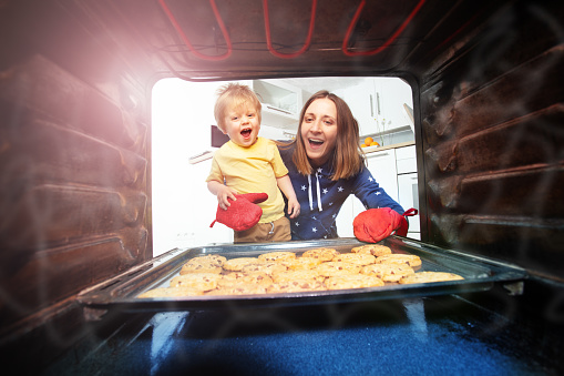 Laughing mother and little boy with oven-gloves take cookies tray from the oven, view from inside