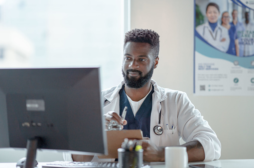 A male doctor of African decent sits behind his desk as he meets virtually online with a patient.  He is looking at his computer screen while simultaneously holding a clipboard and taking notes from the appointment.