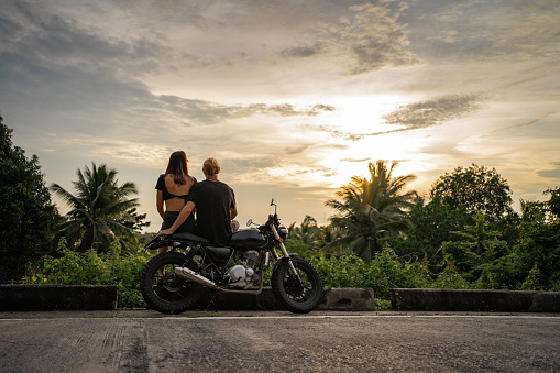 Rear view of couple sitting on a motorcycle by the road and watching amazing sunset.