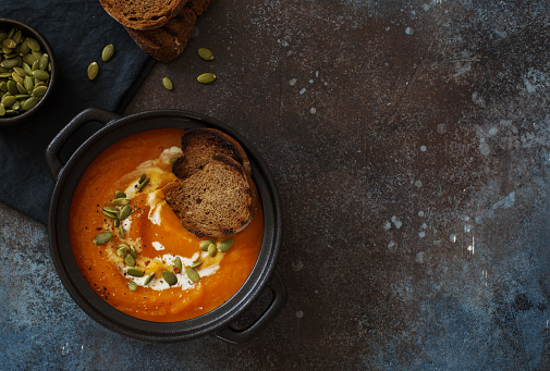 Creamy pumpkin carrots soup with pumpkin seeds and croutons  in black pot on dark stone background. Flat lay. Copy space