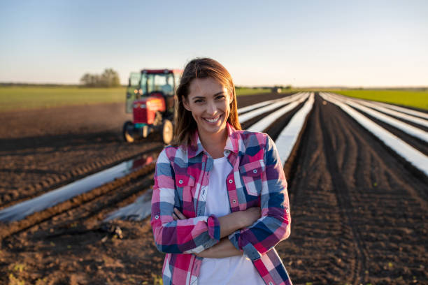 Farmer woman standing with crossed arms in field Female farmer standing with arms crossed looking at camera. Young farmer standing in field with plant foil and tractor smiling. farmer stock pictures, royalty-free photos & images