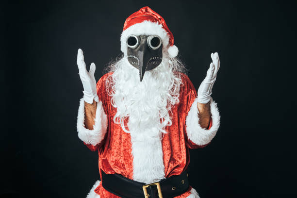 Man dressed as Santa Claus with a Victorian bubonic plague mask, raising hands, on black background. Concept of Christmas, Santa Claus, gifts, celebration. stock photo