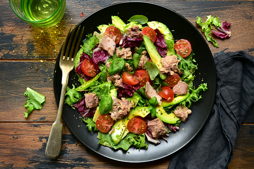 Tuna salad with tomatoes and avocado on a black plate over dark wooden background. Top view with copy space.