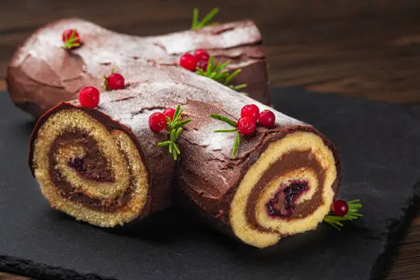 Traditional homemade Christmas cake. Yule log or Buche de Noel. Sponge cake with chocolate cream, ganache, decorated with cranberries Dark wooden background.