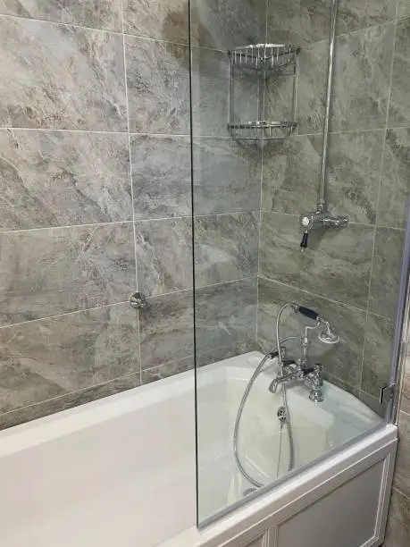 Stock photo showing luxury bathroom suite with white and grey marble wall and floor tiles, white ceramic sink, WC toilet, bath with stainless steel Victorian mixer tap and shower plumbing, artificial plants, mirror and towel rail.