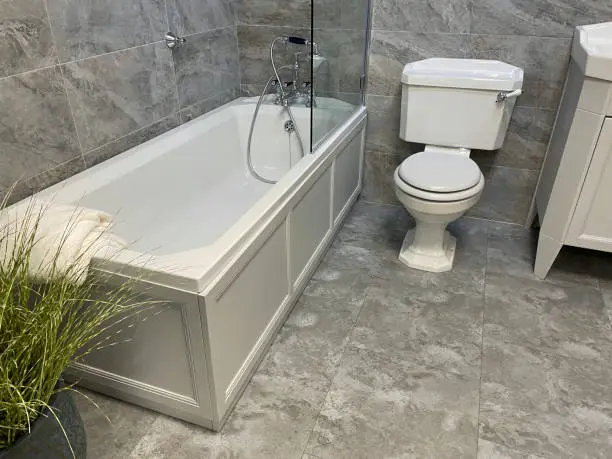 Stock photo showing luxury bathroom suite with white and grey marble wall and floor tiles, white ceramic sink, WC toilet, bath with stainless steel Victorian mixer tap and shower plumbing, artificial plants, mirror and towel rail.