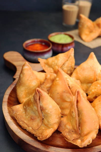 Close-up image of wooden chopping board full of fried samosas, stuffed with spiced potato, peas and meat, mint coriander dip and mango chutney ramekins, drinking glasses of chai tea, focus on foreground stock photo