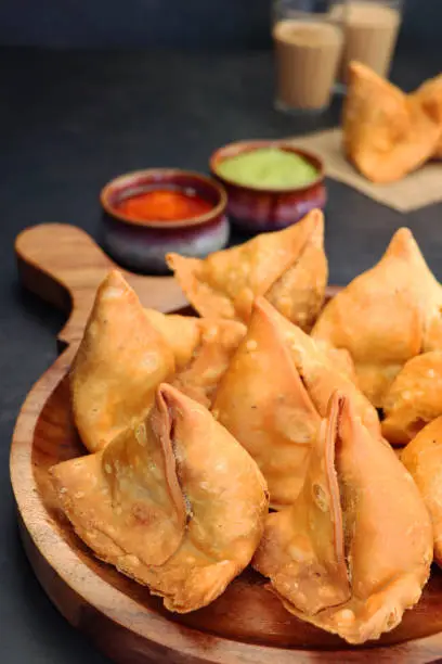 Photo of Close-up image of wooden chopping board full of fried samosas, stuffed with spiced potato, peas and meat, mint coriander dip and mango chutney ramekins, drinking glasses of chai tea, focus on foreground