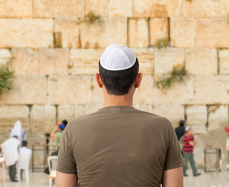 Back of a young tourist in casual clothes and kippah on the head looking at the wailing wall and religious praying near the sacred place of Judaism
