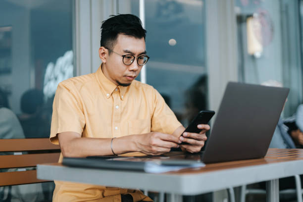 istock Confident young Asian businessman sitting outdoors in a cafe, using smartphone while working on laptop. Business on the go concept 1350394456
