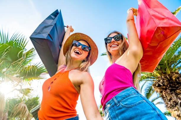 zwei schöne junge frauen, die gerne auf der straße der stadt einkaufen -glückliche und lächelnde schwestern nach dem einkaufen heben ihre einkaufstaschen in den himmel - sale, consumerism and people concept - kaufsucht stock-fotos und bilder