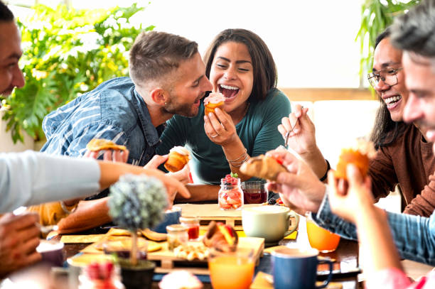 friends having fun at breakfast and eating muffins at bakery or pastry shop - beautiful happy couple eating cupcake with whipped cream in a cafe - young woman is feeding her boyfriend - eating people group of people home interior imagens e fotografias de stock