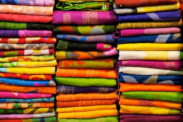 Artistic variety shade tone colors ornaments patterns, closeup view of stacked saris or sarees in display of retail shop.