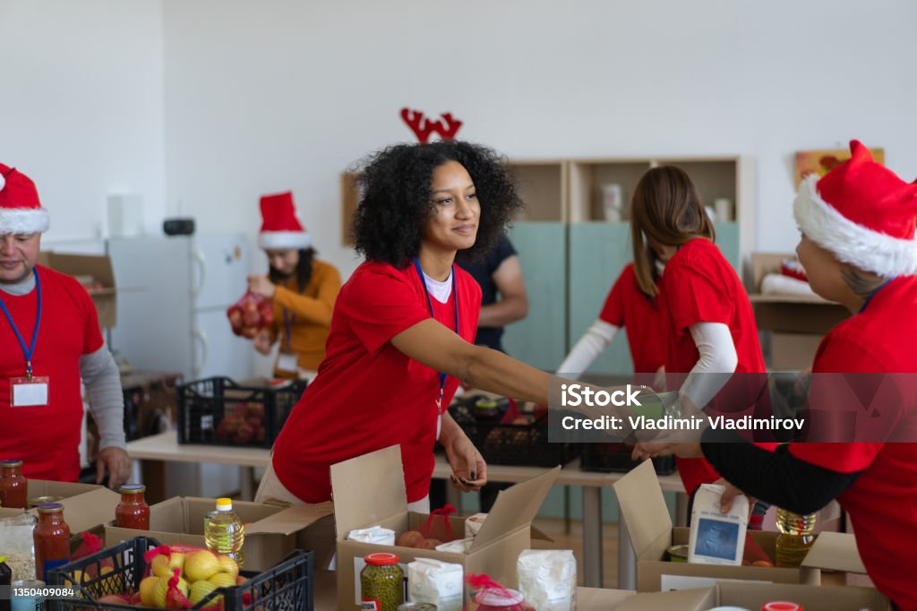 Volunteers distributing grocery donations Smiling helpers assisting each other in organising food donations, packing the groceries into cardboard boxes, wearing red t-shirts and Santa hats for Christmas spirit Christmas Stock Photo