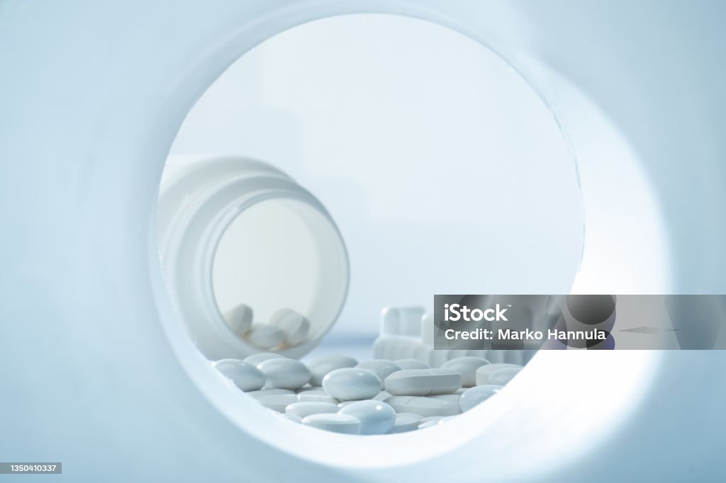 Closeup of white pills against white background with plastic pill container in the background with plastic pill container Helsinki / Finland - OCTOBER 31, 2021:Closeup of white pills against white background with plastic pill container in the background with plastic pill container Pharmacy Stock Photo