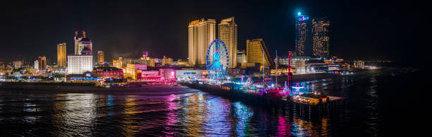 Panoramic aerial view of the Broadwalk on the waterfront in Atlantic City Downtown, the famous gambling center of the East Coast USA, with multiple casinos and amusing park with a Ferris Wheel on a pier. Night view of the Broadwalk along the Downtown Atlantic City's waterfront, New Jersey, USA. Extra-large high-resolution aerial stitched panorama. boardwalk stock pictures, royalty-free photos & images