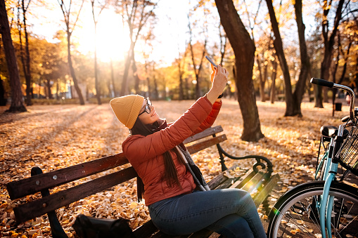 Woman sitting on the bench at autumn city park and using a smartphone
