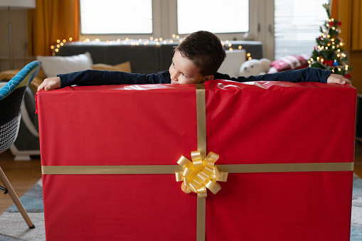 A young joyous boy is stretching both arms out to try and touch both ends of a very large red-wrapped gift at Christmas time