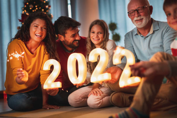 Family holding illuminative numbers 2022 while celebrating New Year Happy family celebrating New Years Eve at home with kids, sitting by the Christmas tree, holding sparklers and illuminative numbers 2022 representing the upcoming New Year 2022 photos stock pictures, royalty-free photos & images