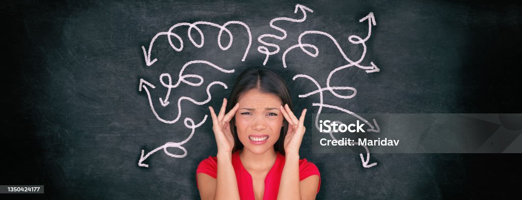 Confused woman confusion illustrated on blackboard with chalk drawing of arrows going everywhere around head showing indecision. Indecisive stressed Asian girl with headache panoramic banner. Chaos Stock Photo