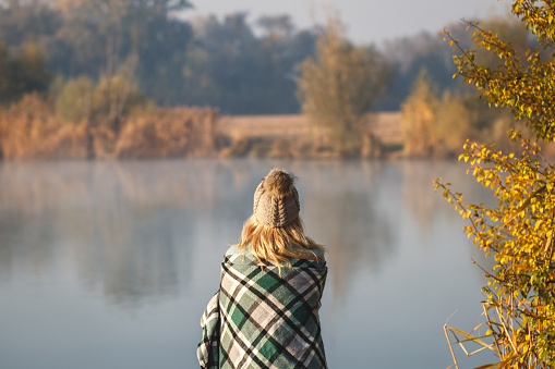 Female person relaxing in cold autumn morning in nature. Camping outdoors. Woman with knit hat is wrapped in warm blanket at lake.