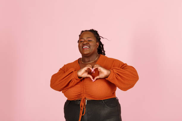 Smiling young black plus size woman shows heart with palms on pink background stock photo