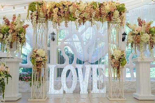 Beautiful white wedding arch for wedding ceremony decorated with flowers, lightbulbs and sign Love with big white letters, copy space