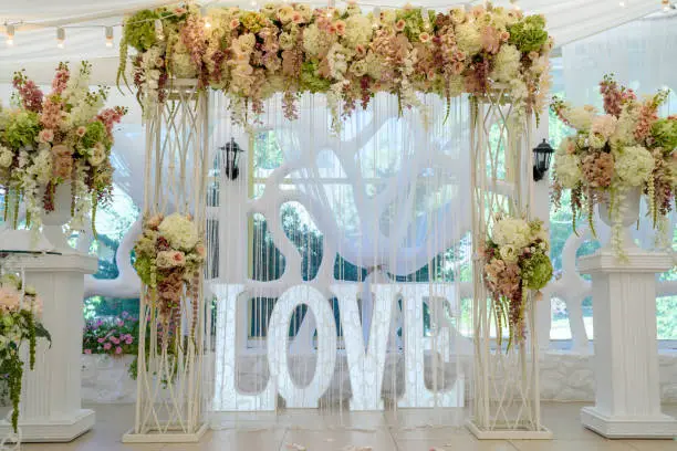 Photo of Beautiful white wedding arch for wedding ceremony decorated with flowers, lightbulbs and sign Love with big white letters, copy space