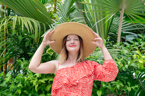 Beautiful young woman wearing an off the shoulder summer dress and a large straw hat, while holding her eyes closed, smiling and enjoying the surrounding tropical environment with lush exotic trees.