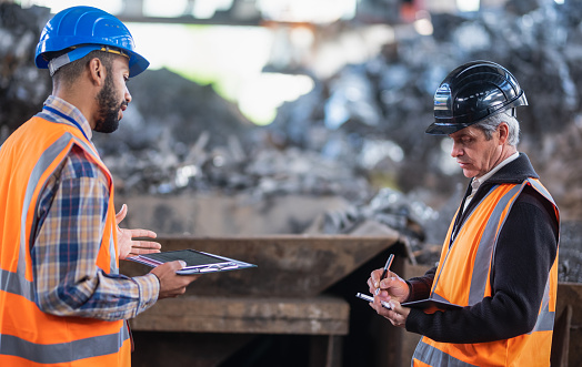 Side View Of Two Colleagues In Safety Reflective Vests Standing In Factory In Front Of Pile Of Scrap Metal