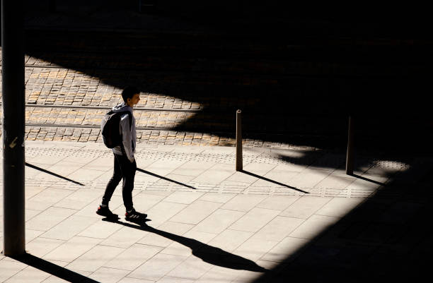One teenage boy standing alone waiting for a tram on bus stop in sunlight, with shadows stock photo