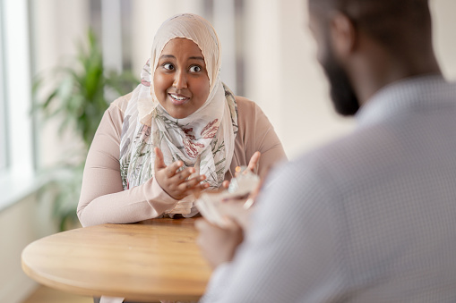 A Muslim patient has an puzzled expression on her face as she shares during a therapy session and asks her Therapist a question about her struggle.  She is dressed casually and is wearing a Hijab.  Her male therapist is sitting across from her listening attentively, and taking notes on his clipboard.