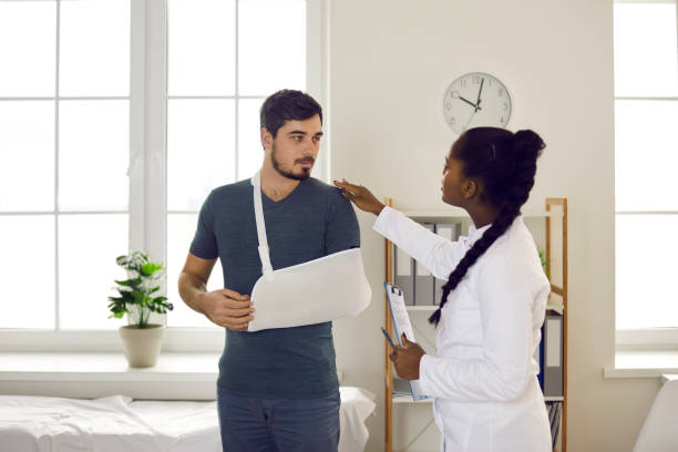African american doctor conduct medical examination on patient with hand injury Traumatology and medicine. African american woman doctor orthopedist physiotherapist conducting medical examination on caucasian man patient with broken hand injury in sling at hospital office ward man with arm sling stock pictures, royalty-free photos & images