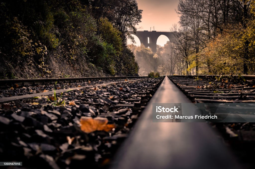 Autumnal view of the Elstertal bridge (German: Elstertalbrücke), a railway bridge near Plauen (Saxony, Germany), built in 1851, today the second largest brick bridge in the world. View along a railway track, in the background sunbeams fall through the early morning fog onto a historic railway viaduct and autumn-colored trees. Journey Stock Photo