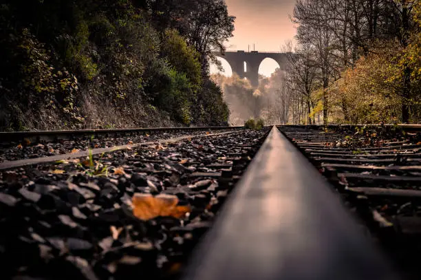 View along a railway track, in the background sunbeams fall through the early morning fog onto a historic railway viaduct and autumn-colored trees.