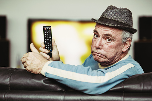 Man in his late 60s grimacing as he sits on a leather couch in front of a large flat-screen TV, holding the remote control.