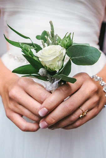 Beautiful bride in white dress holding wedding boutonniere with white rose for groom in hands
