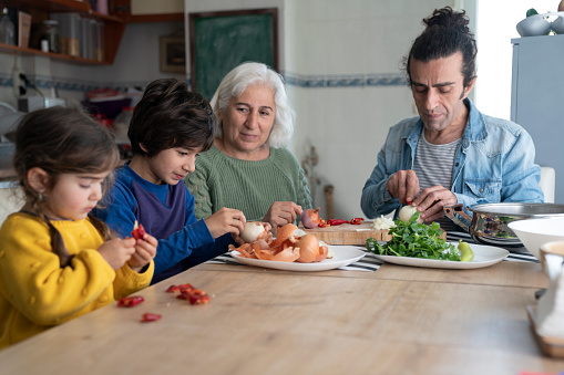 Photo of senior grandmother, mature adult father, 2,5 years old granddaughter and 8 years old grandson preparing food on table in domestic kitchen. Onions and peppers are seen on table. Senior woman has white hair. Shot indoor with a full frame mirrorless camera.
