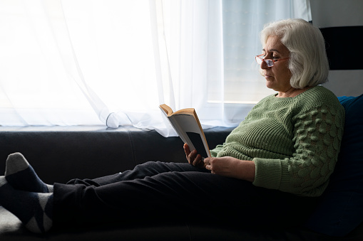 Portrait of senior woman with gray hair lying on sofa in living room and reading book. She is next to window. Shot indoor under daylight with a full frame mirrorless camera.