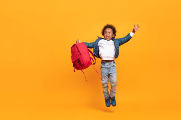 bambino nero bambino 3 anni, studente bambino che salta per godere felice tornare a scuola con borsa di scuola rossa, ritratto isolato su sfondo giallo con spazio di copia, felicità studentesca internazionale - primary colours foto e immagini stock