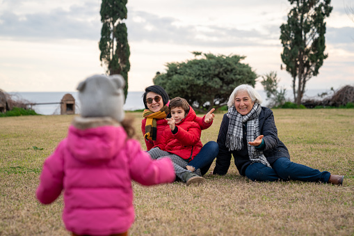 Photo of senior grandmother, mother and son watching baby girl's first steps in public park. baby girl is wearing a pink overcoat. Shot in outdoor public park with a full frame mirrorless camera.