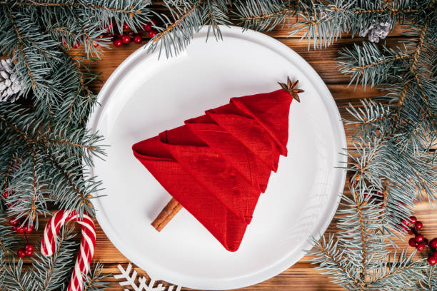 Linen napkin folded in shape of Christmas tree on white plate among spruce branches. New Year table setting. Top view stock photo