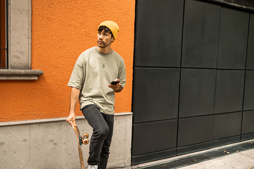 Front view on a young Latin with the skateboard in the hand. He is using mobile phone.