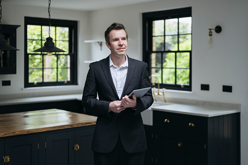 Portrait of mid adult Caucasian man in full suit holding digital tablet and looking away from camera with kitchen island and granite countertops in background.