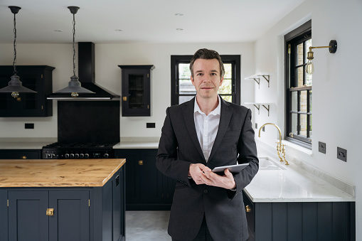 Front view of confident mid 30s real estate agent holding digital tablet and looking at camera with luxurious kitchen features in background.