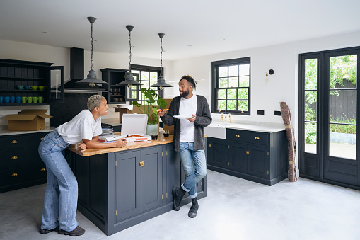 Full length view of casually dressed man and woman in their 30s eating and talking in between moving belongings into their new Shaker-style kitchen.