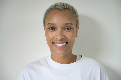 Head and shoulders view of woman with short, bleached, natural hair wearing white t-shirt and smiling at camera against white background.