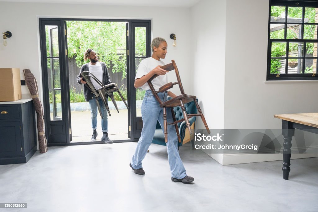 Man and woman bringing chairs into home on moving day Full length view of casually dressed Black couple carrying chairs through patio doors into spacious dining area adjoining kitchen. Chair Stock Photo
